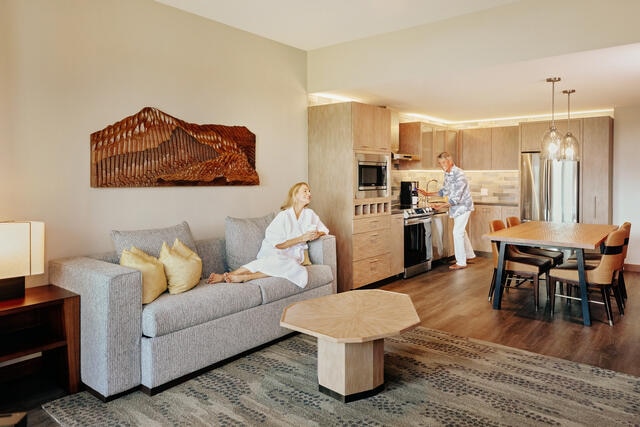 Mature woman enjoying coffee on the couch,while mature man prepares food in the in-suite kitchen, Maui Bay Villas, a HIlton Grand Vacations Club, Hawaii. 