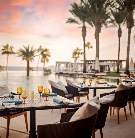 Tables and chairs at an outdoor restaurant by the ocean with palm trees
